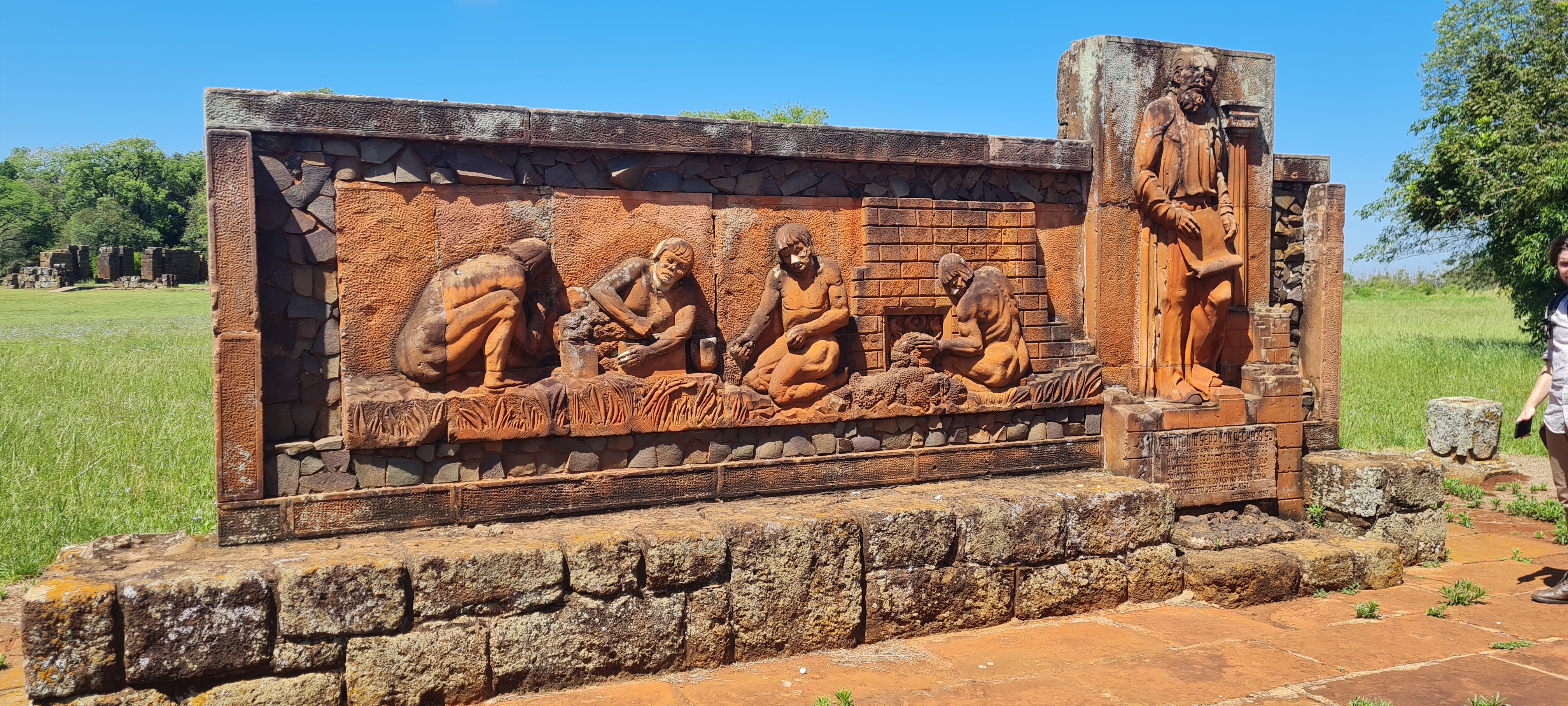 Monumento em homenagem aos indígenas siderúrgicos no Sítio Arqueológico de São João Batista, Rota das Missões, Rio Grande do Sul, Brasil A fotografia colorida mostra uma pedra alaranjada com entalhes de indígenas trabalhando com ferro ao lado de um padre
