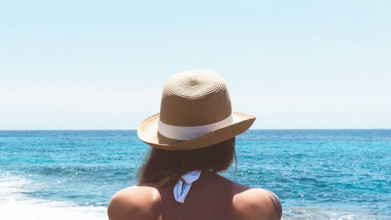 Mulher de chapéu, de costas, olha para o mar azul em praia