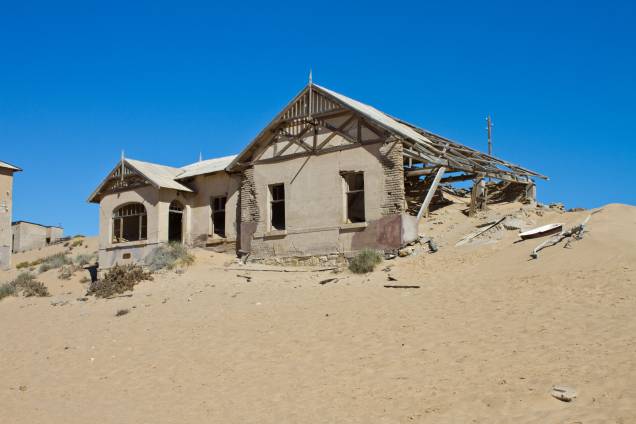<strong>Cidade Fantasma de Kolmanskop, Namíbia</strong> Os desertos africanos possuem um fascínio que desperta a curiosidade dos visitantes, sobretudo o Mar de Areia da Namíbia, tombado pela Unesco em 2013 como Patrimônio Mundial. Foi a areia, aliás, a responsável por engolir as casas construídas na cidade de Kolmanskop. No início de 1900, a região foi ocupada pelos alemães, que viram nela a chance de enriquecer graças à exploração de diamantes. O excesso da atividade, no entanto, fez com que os minérios sofressem um forte esgotamento. Na década de 1950, o abandono por parte de seus moradores fez com que a constante limpeza da areia insistente que ocupava as suas ruas fosse completamente abandonada. Hoje, o governo mantém a região habitável para que os turistas curiosos que passeiam por ali possam continuar a explorar a área <strong>Cidade Fantasma de Kolmanskop, Namíbia</strong> Os desertos africanos possuem um fascínio que desperta a curiosidade dos visitantes, sobretudo o Mar de Areia da Namíbia, tombado pela Unesco em 2013 como Patrimônio Mundial. Foi a areia, aliás, a responsável por engolir as casas construídas na cidade de Kolmanskop. No início de 1900, a região foi ocupada pelos alemães, que viram nela a chance de enriquecer graças à exploração de diamantes. O excesso da atividade, no entanto, fez com que os minérios sofressem um forte esgotamento. Na década de 1950, o abandono por parte de seus moradores fez com que a constante limpeza da areia insistente que ocupava as suas ruas fosse completamente abandonada. Hoje, o governo mantém a região habitável para que os turistas curiosos que passeiam por ali possam continuar a explorar a área