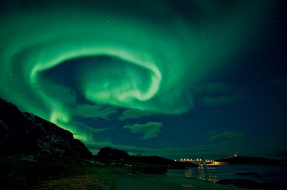 Uma aurora paira sobre a ponte Sommarøy, na ilha Kvaløy, no norte da Noruega, em uma semana de atividade solar intensa. As auroras são causadas pelo impacto de partículas carregadas nos gasesda atmosfera. Mais comuns perto dos polos, as auroras também ocorremem latitudes mais baixas durante tempestades solares fortes. Uma aurora paira sobre a ponte Sommarøy, na ilha Kvaløy, no norte da Noruega, em uma semana de atividade solar intensa. As auroras são causadas pelo impacto de partículas carregadas nos gasesda atmosfera. Mais comuns perto dos polos, as auroras também ocorremem latitudes mais baixas durante tempestades solares fortes.