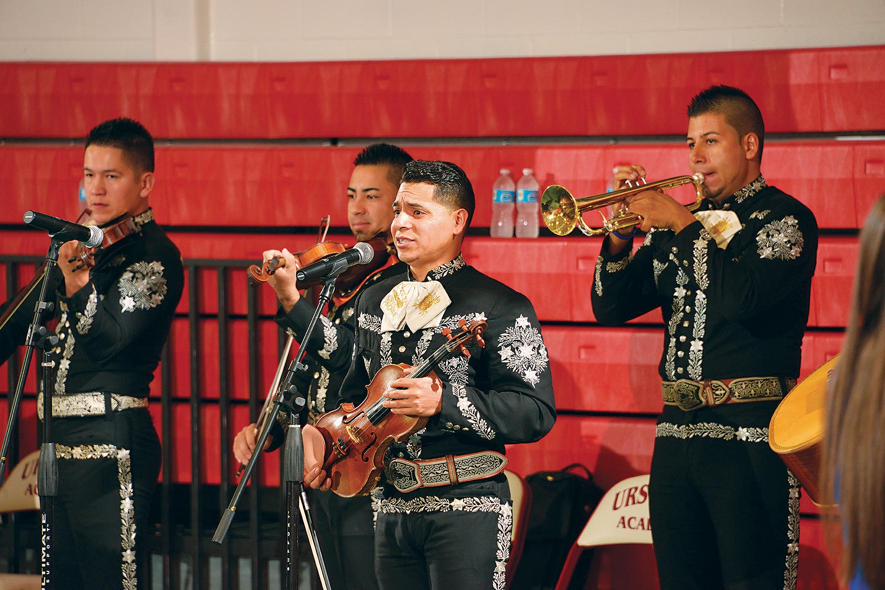 Quatro m&uacute;sicos, tr&ecirc;s segurando violinos e o quarto um trompete, tocam e cantam para uma plateia, vestidos com fardas t&iacute;picas pretas com detalhes em branco