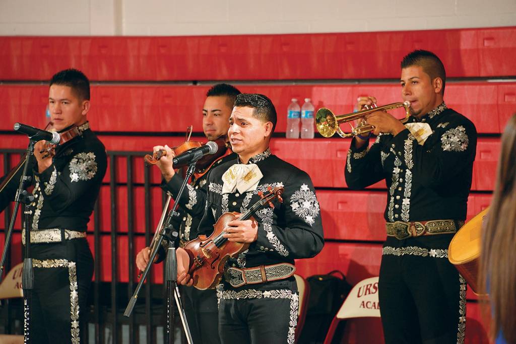 Quatro músicos, três segurando violinos e o quarto um trompete, tocam e cantam para uma plateia, vestidos com fardas típicas pretas com detalhes em branco