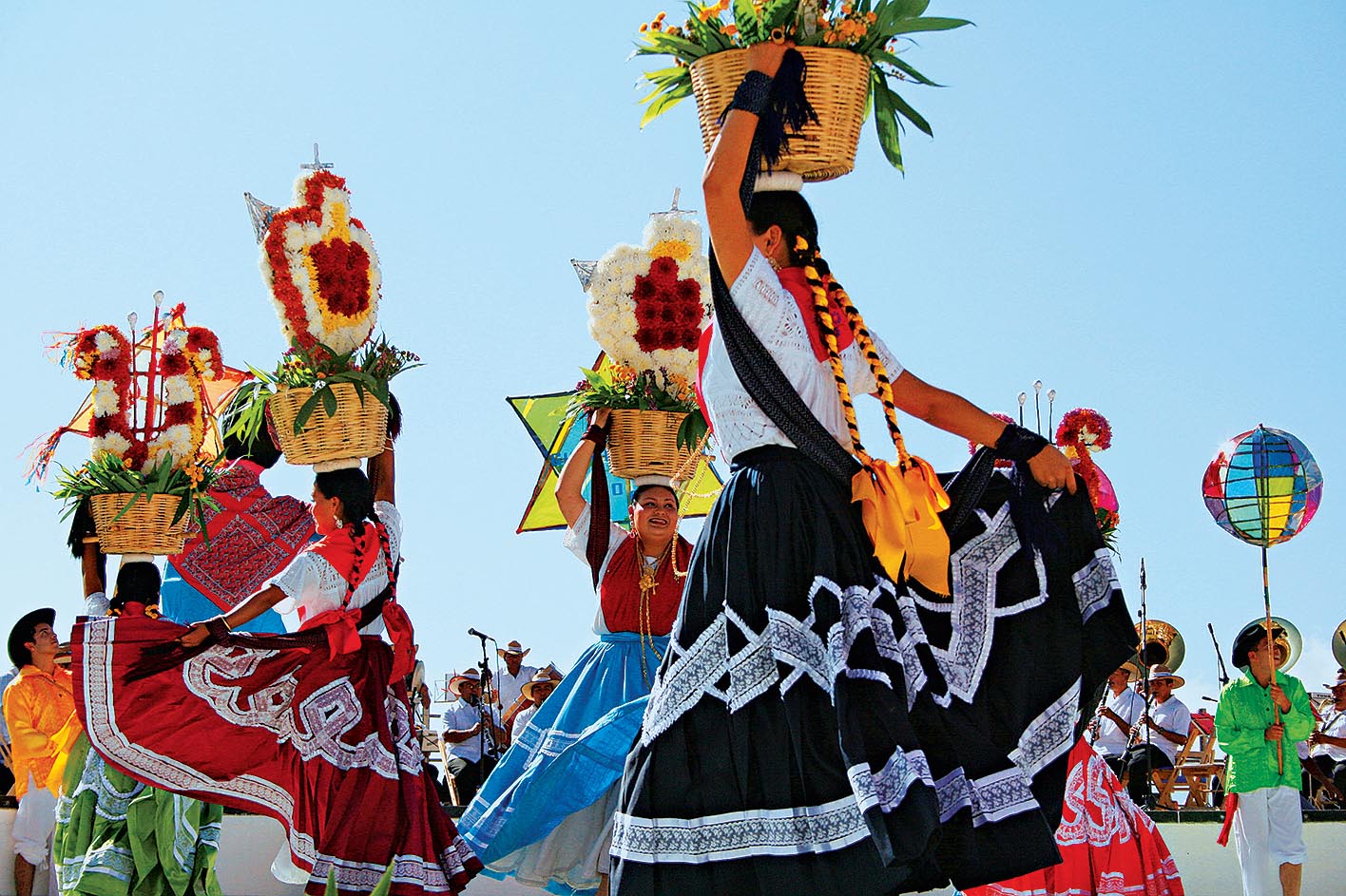 Mulheres dan&ccedil;am ao som de um grupo musical ao vivo, com longas tran&ccedil;as e saias compridas. Em uma m&atilde;o, seguram uma cesta de flores apoiada na cabe&ccedil;a, enquanto com a outra levantam uma ponta da saia para dan&ccedil;ar