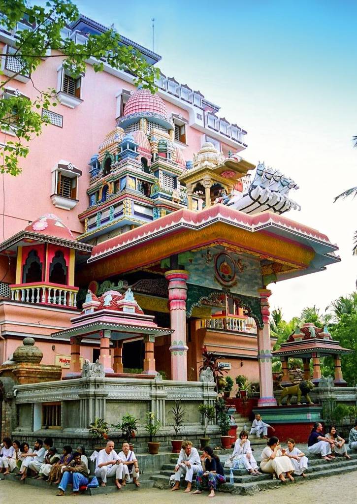 Templo, Amritapuri, Índia Um templo alto, diversas ramificações verticais e esculturas de cavalos empinados na parte de cima da entrada. Um número de pessoas se senta nos degraus do templo, ocupando-os quase inteiros.