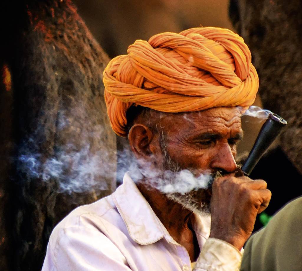 Homem de turbante fuma chilam, soltando uma trilha de fumaça de sua boca