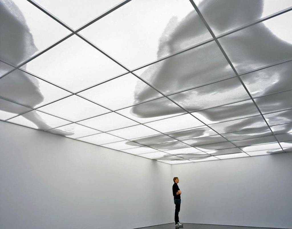 Uma pessoa, sozinha, se encontra em um quarto desprovido de móveis, com um teto onde sombras se movem, tomando a forma de nuvens