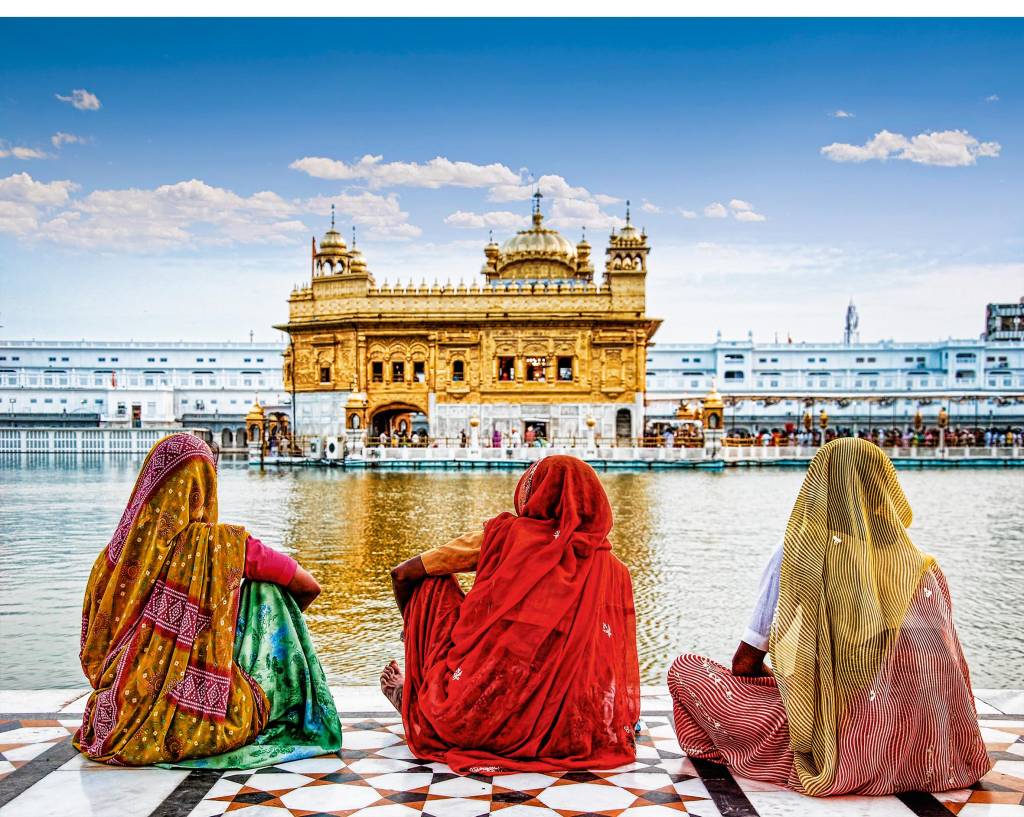 Templo Dourado, Amritsar, Índia Três mulheres vestidas em sáris sentam-se à beira do rio, observando a movimentação na outra margem, em volta do Templo Dourado.