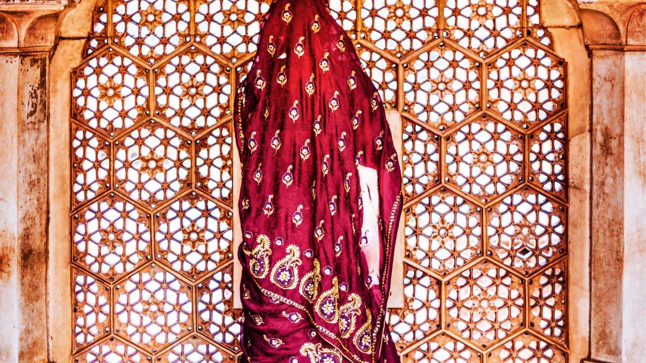 Mulher vestida em um sári se posiciona de frente a uma janela, cuja abertura está divida em furos formados por várias mandalas esculpidas