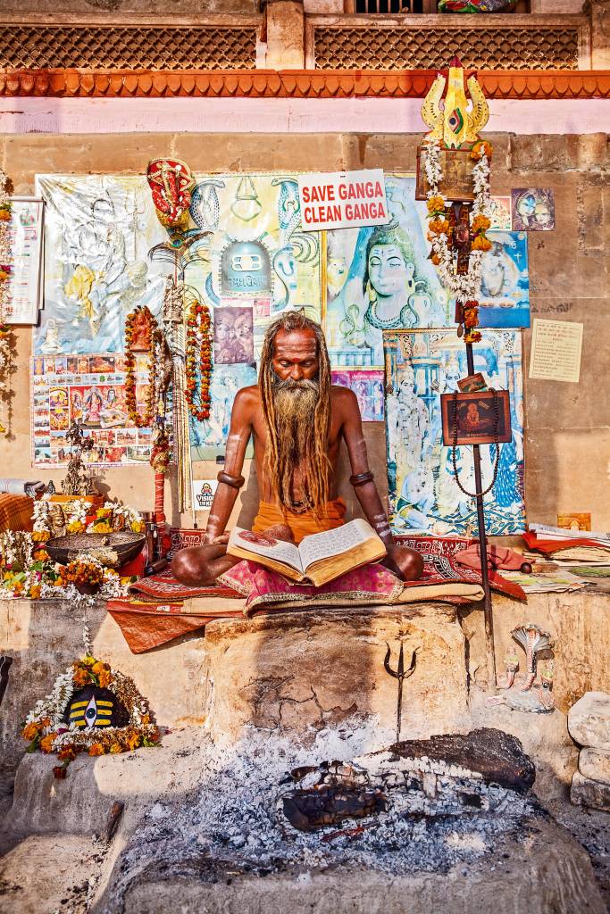 Sadhu, Varanasi, Índia Sadhu está sentado em meio a múltiplos pôsteres e lembrancinhas, lendo um livro em pose de disciplina
