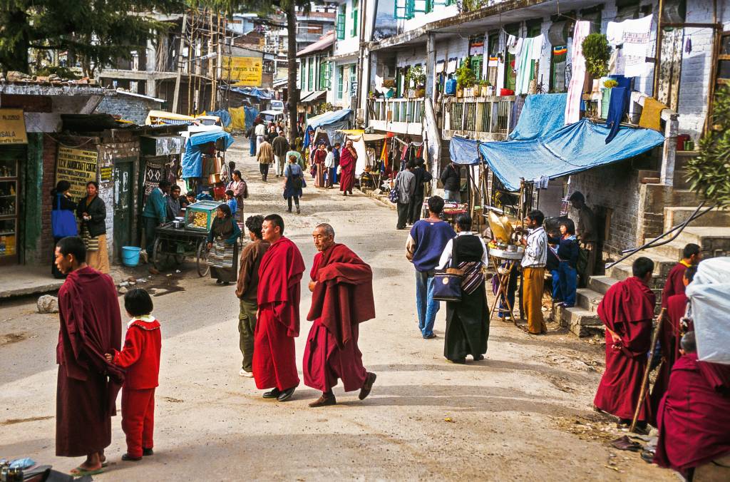 Budismo, Dharamshala, Índia Pedestres e monges circulam por uma rua de terra, com alguma tendas de comércio instaladas esparsamente