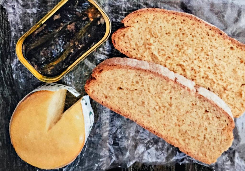 Duas fatias de pão, uma lata de sardinhas e uma roda de queijo do tamanho da lata, sobre um fundo de papel plástico