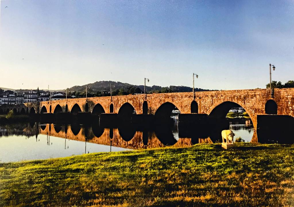 Dividindo a imagem em dois, a ponte de Lima, formada por diversos arcos com bases submersas, parece se aproximar do leitor graças ao ângulo lateral de fotografia. Do lado de cá, grama; do lado de lá, alguns prédios antigos.