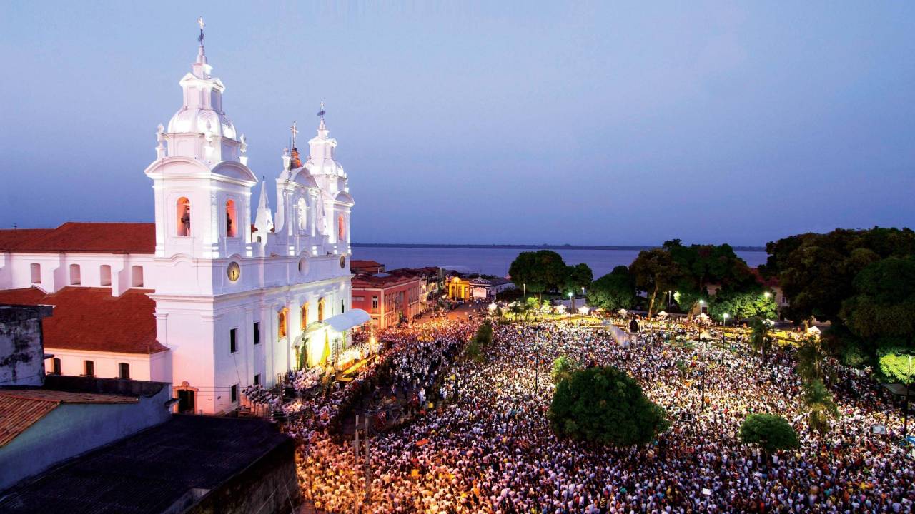 Enorme catedral, no crepúsculo, com a fachada iluminada. Em frente, uma multidão de pessoas se reúne em procissão, ocupando a parte inferior inteira da foto.