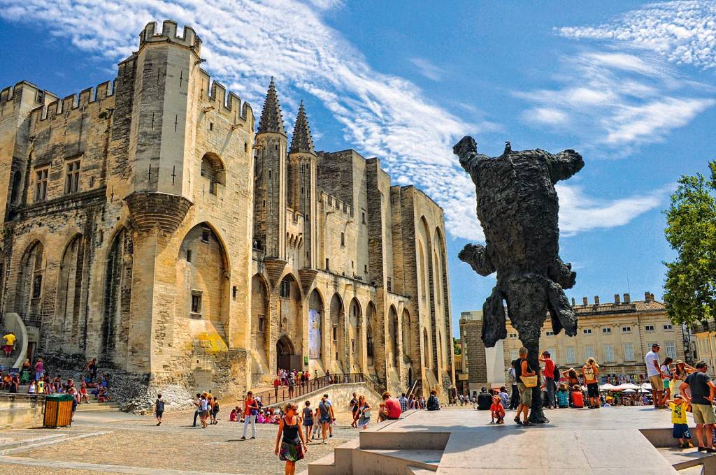 Um palácio medieval imponente se ergue no canto esquerdo da foto, cercado por turistas. À direita, uma escultura imita um elefante se equilibrando perfeitamente pela tromba