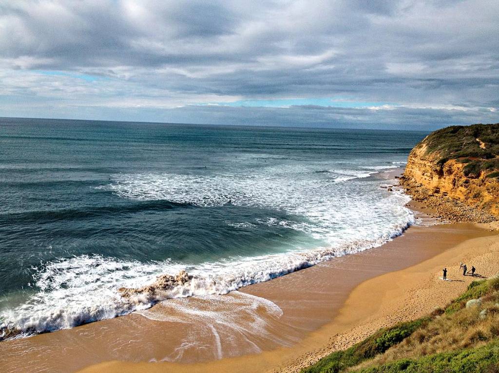 Ondas quebram em uma praia, enquanto surfistas preparam pranchas de surfe. O local é emoldurado por uma elevação, de onde a foto é tirada. Ou seja, toda a cena é vista do alto.