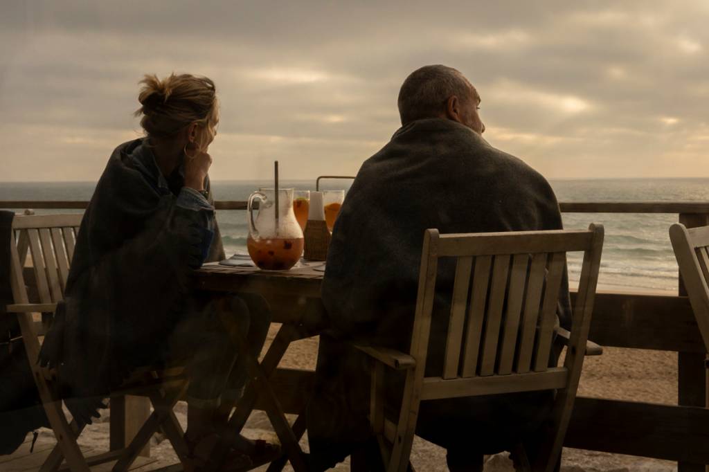 A imagem mostra um casal de costas para a câmera. Eles estão sentados à mesa, de frente para o mar, conversando e bebendo suco de laranja.