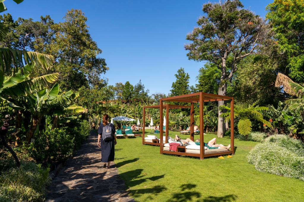 Mulher com mochila nas costas caminha pelo gramado cercado de árvores onde estão day beds e espreguiçadeiras com pessoas tomando sol