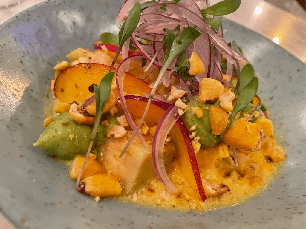 Prato de cerâmica cinza com cubos de peixe, fatias de nectarina, cebola roxa, milho frito e um caldo amarelo ao fundo