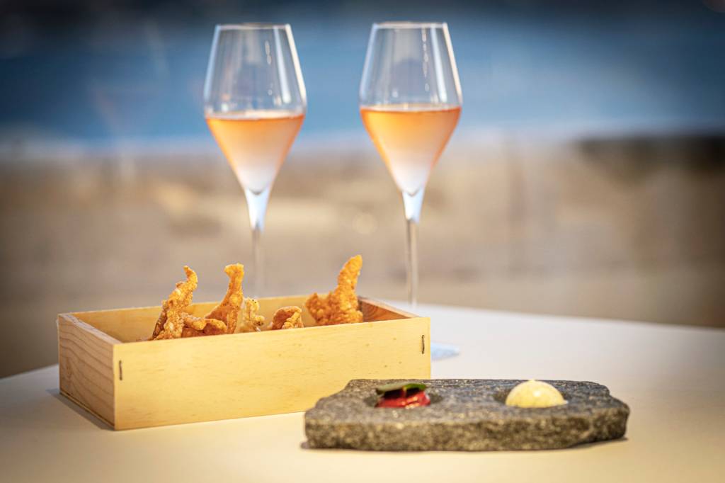 Duas taças de espumante rosé ao fundo e, em primeiro plano, um prato de pedra cinza com manteigas e uma caixa de madeira com crisps de arroz