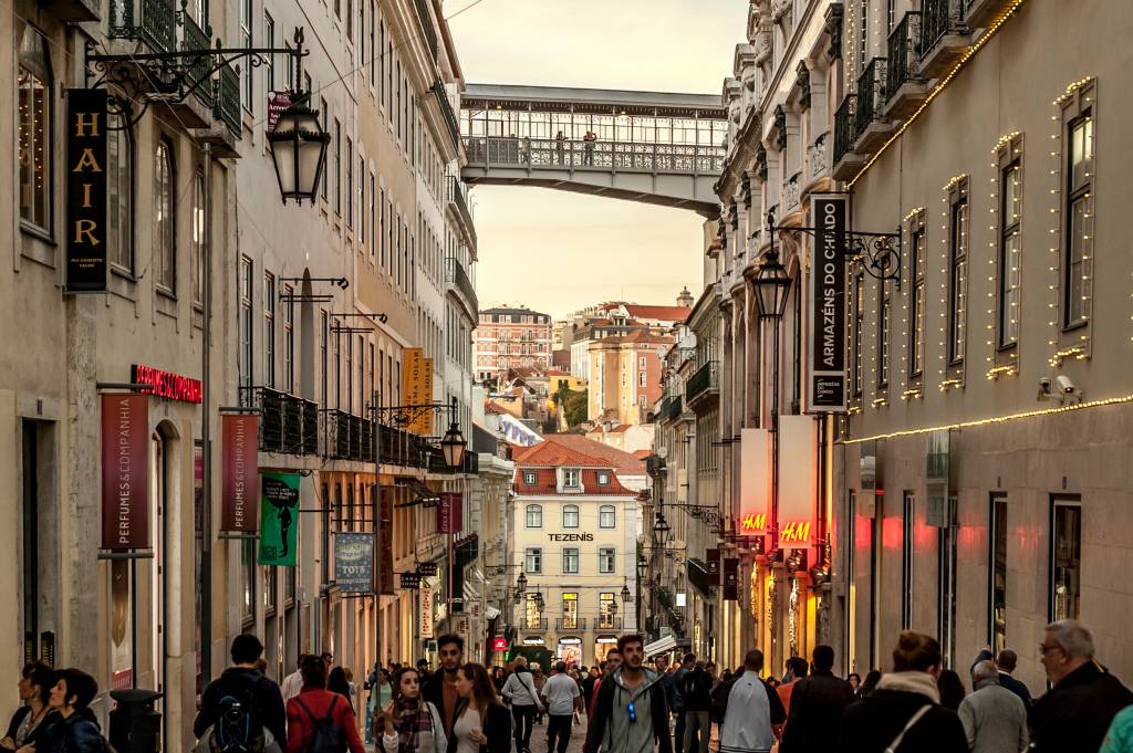 Rua cheia de pessoas com construções antigas dos dois lados e uma ponte de ferro ligando os dois lados