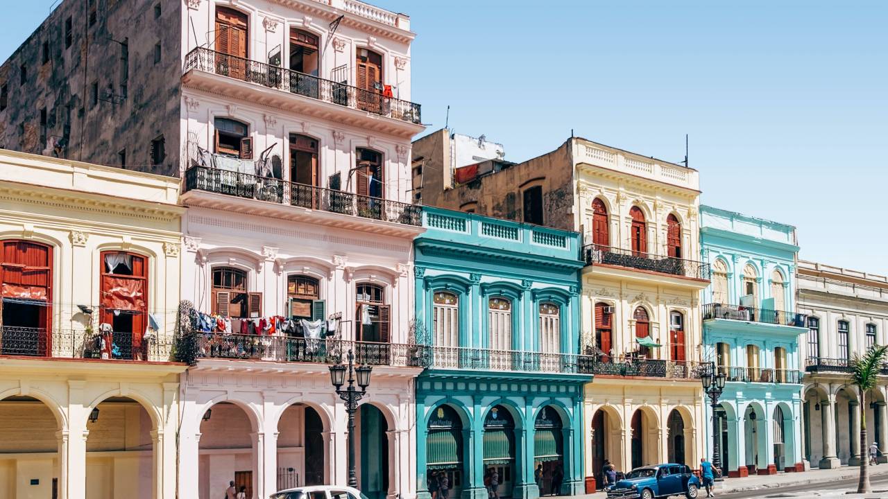 A imagem mostra construções antigas de vários andares e de várias cores diferentes. Na rua, em frente a construção, há um carro antigo de cor verde água.