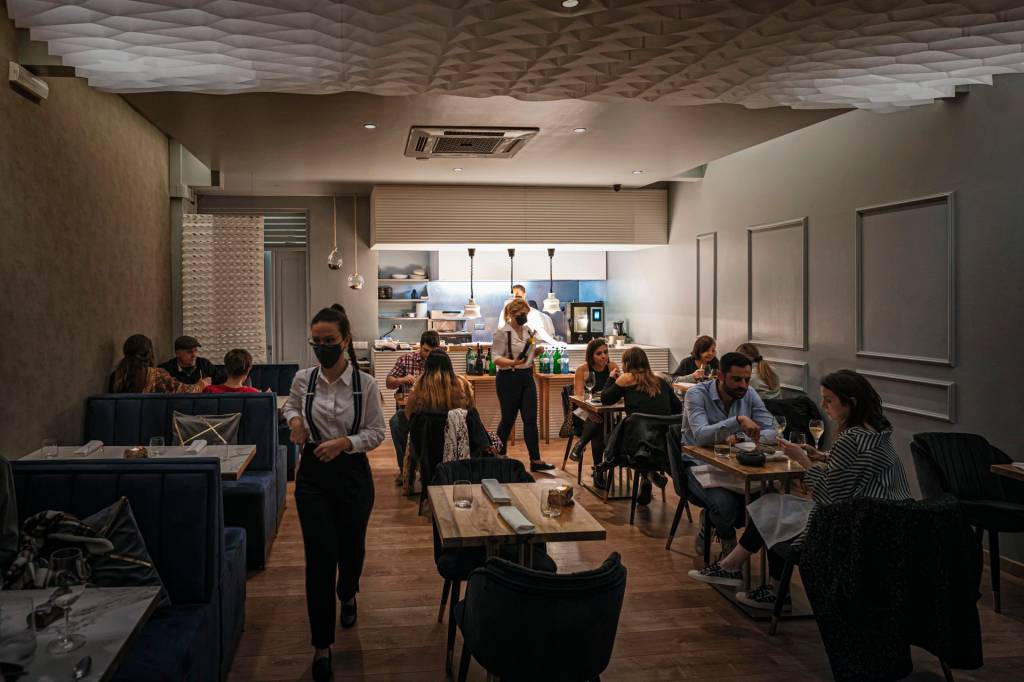 Sala de restaurante com algumas mesas de madeira ocupadas, garçons circulando entre elas e a cozinha aberta ao fundo