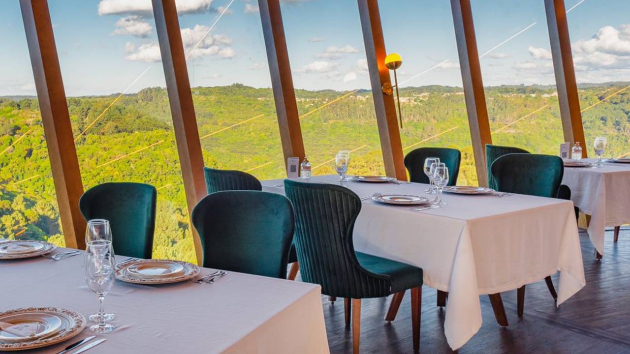 Na imagem vemos o interior de um restaurante. Há várias mesas com toalhas brancas e cadeiras pretas. As paredes do restaurante são feitas de janelas, assim, vemos um vale verde que est[a ao lado do restaurante.