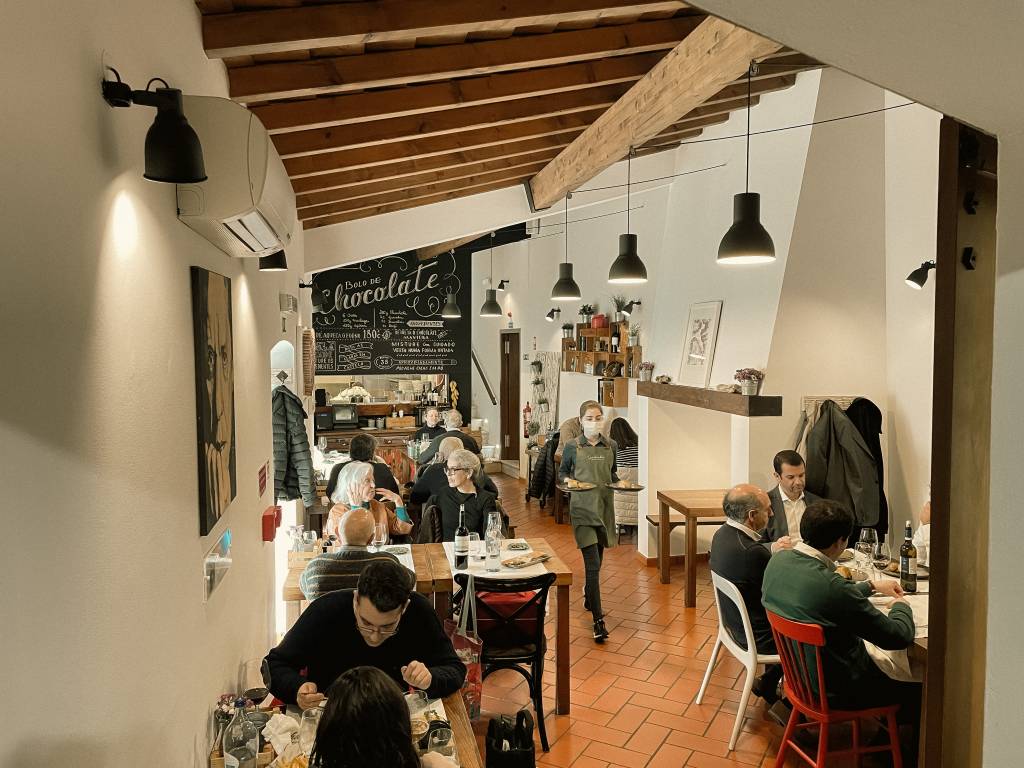 Sala de um restaurante com pessoas sentadas nas mesas e um garõm passando com a bandeja