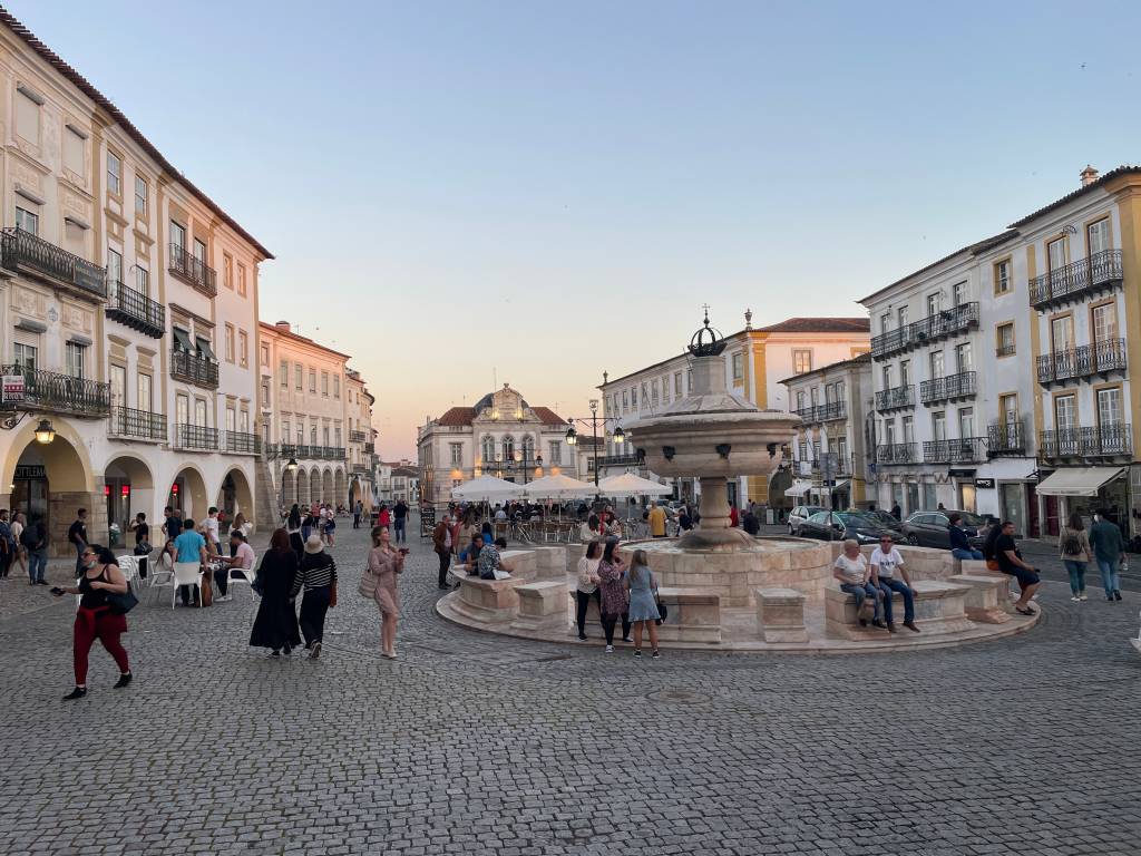 Grande praça calçada de paralelepípedos com uma fonte no centro, cercada de palacetes de dois e três andares e paredes brancas, com pessoas passeando