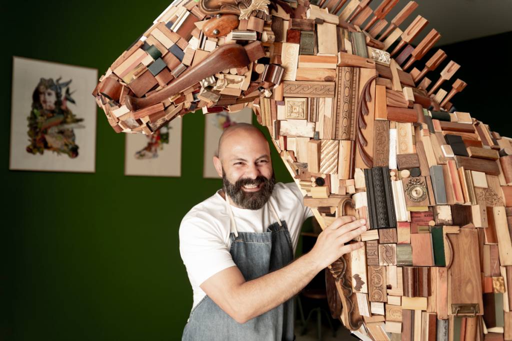 OficinaMarques_GezoMarques_BrunoBarata-2 Artista careca, de barba e bigodes, camiseta branca e avental jeans, posa com a sua escultura de cavalo em tamanho real, feita com pedaços de madeira