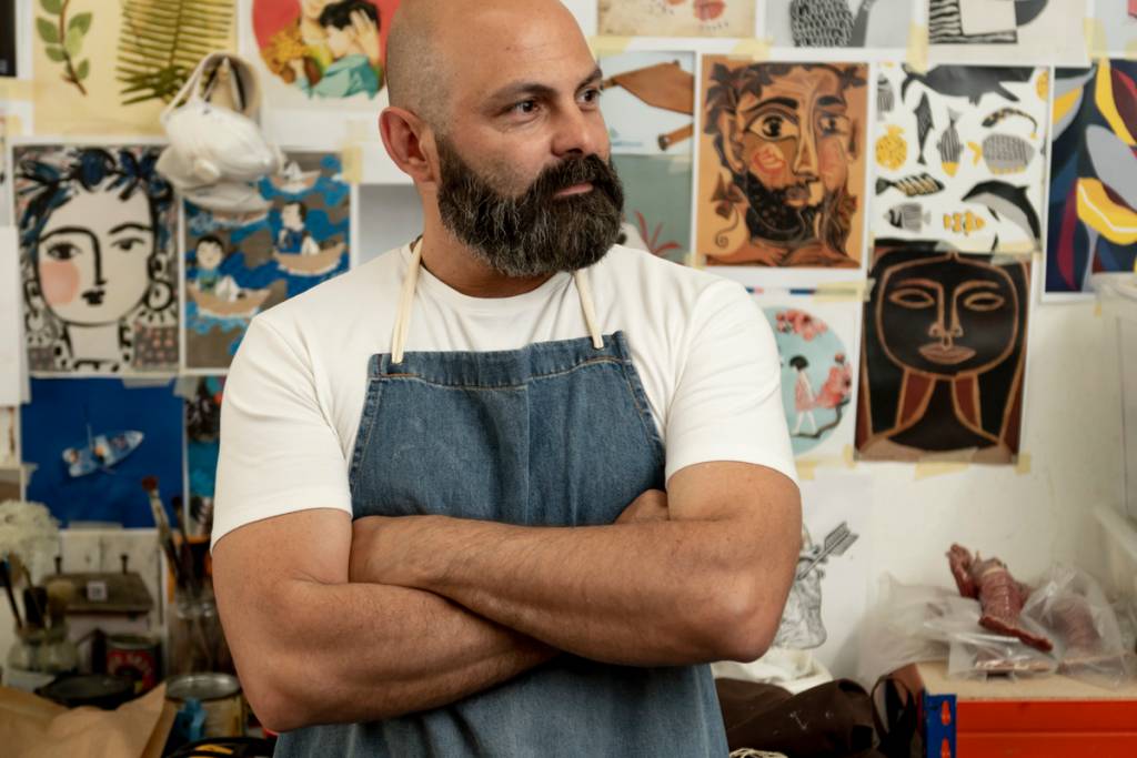 OficinaMarques_GezoMarques_BrunoBarata-5 Artista careca, de barba e bigode, posa em seu ateliê, em frente a uma parede cheia de gravuras coloridas