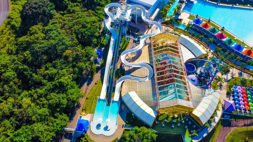 Wet’n Wild – Surge e Bubba Tub A imagem aérea do parque aquático mostra dois toboáguas grandes, um balcão e um outro brinquedo de escorregar, além de mata verde e tendas coloridas.