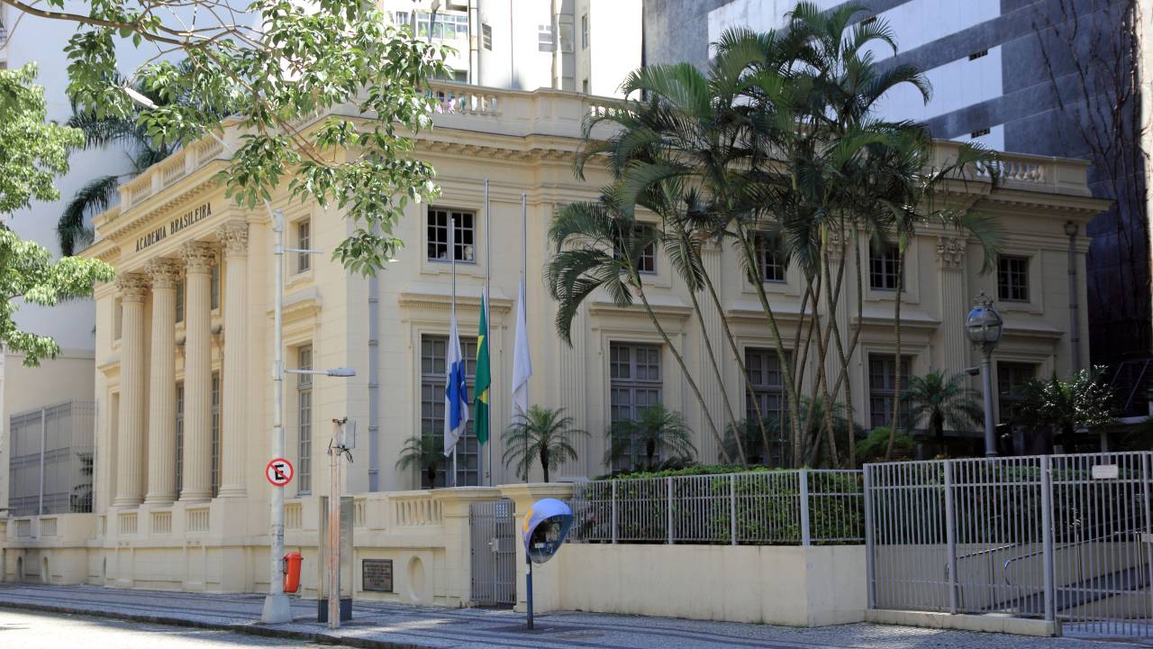 A imagem colorida mostra a lateral e parte da frente de um prédio de cor creme. Ele tem a estrutura quadrada e uma estética clássica