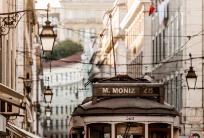 Prédios de fachadas em tons pastel com luminárias e um pedaço de um bonde elétrico com uma placa que diz M Moniz