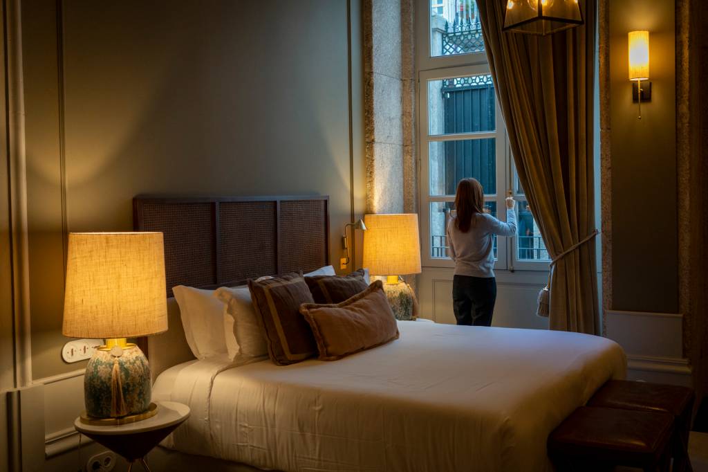 Quarto de hotel com almofadas sobre a cama, abajures acesos nas mesas de cabeceira e uma mulher de costas abrindo a cortina