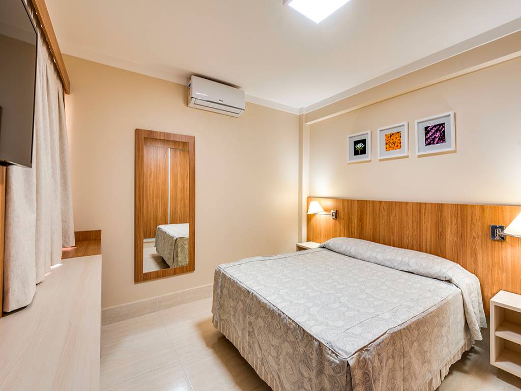 Quarto de hotel com paredes bege, cama de casal com colcha estampada, cabeceira de madeira clara, duas mesas de cabeceira com lumin&aacute;rias, espelho de corpo inteiro, televis&atilde;o na parede e ar condicionado