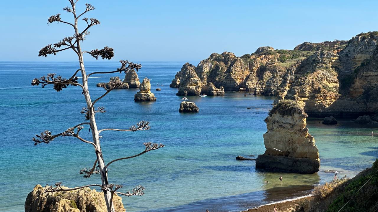 Praia de areas douradas e águas azuis vista do alto das falésias, com grandes rochas dentro do mar