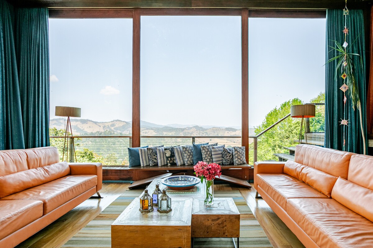 Sala de estar com janelas panor&acirc;micas e vista para montanhas. Dois sof&aacute;s de couro laranja se enfrentam, com uma mesa de centro de madeira r&uacute;stica entre eles, decorada com um vaso de flores vermelhas e lanternas. Cortinas azuis escuras emolduram a vista, e um banco de madeira com almofadas estampadas fica sob a janela. O ch&atilde;o &eacute; de madeira clara com um tapete listrado. Duas lumin&aacute;rias de ch&atilde;o com c&uacute;pulas redondas iluminam o ambiente. Uma escada de madeira com corrim&atilde;o met&aacute;lico &eacute; vis&iacute;vel &agrave; direita, levando a um andar superior. A paisagem exterior &eacute; de colinas verdes sob um c&eacute;u azul claro.