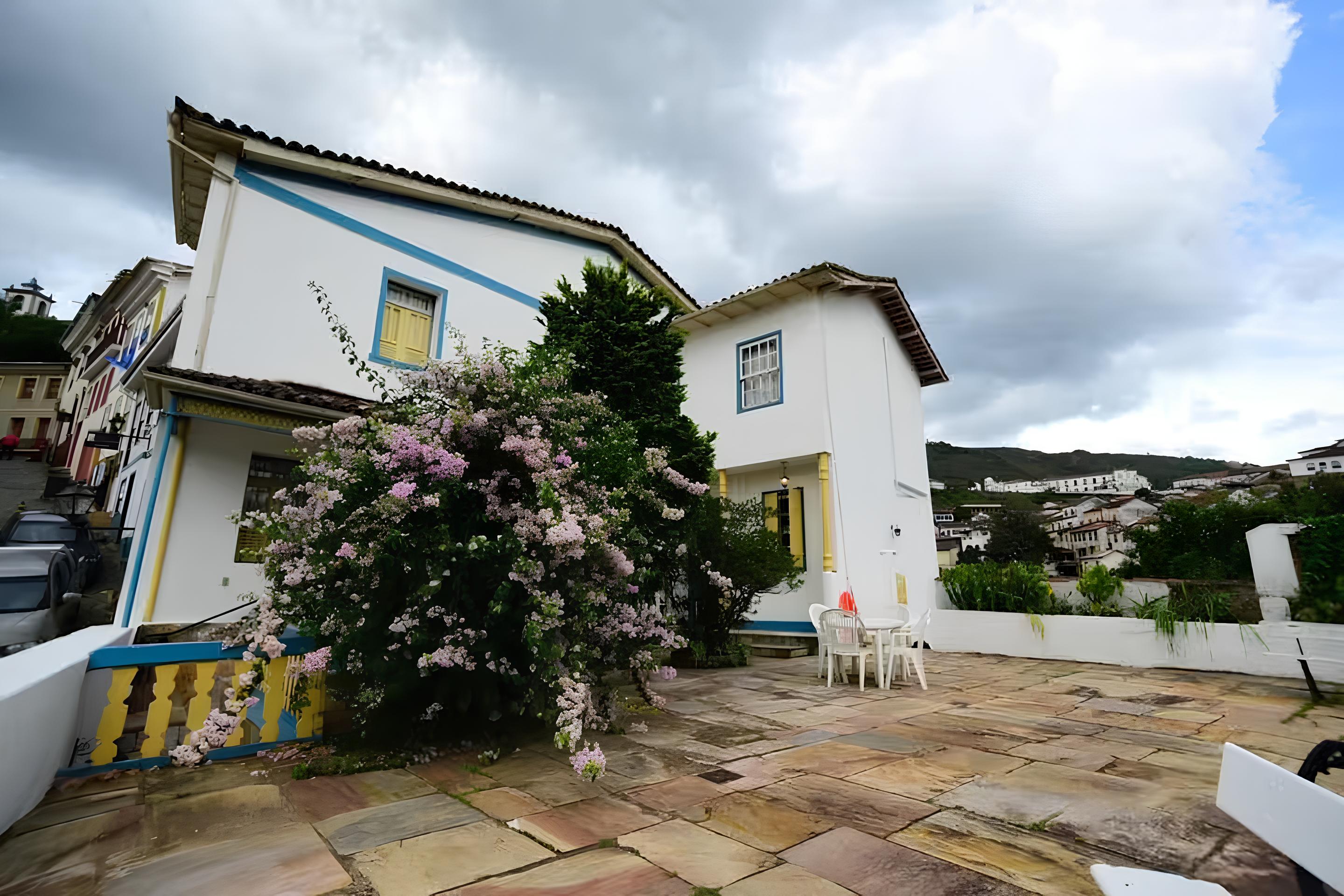 Casas brancas com detalhes azuis e amarelos em Ouro Preto, Minas Gerais. Um arbusto florido em tons de rosa e roxo se destaca no centro. &Agrave; direita, um p&aacute;tio de pedras com mesa e cadeiras brancas. Ao fundo, mais casas e montanhas sob c&eacute;u nublado