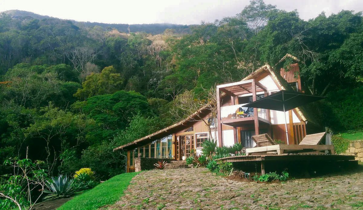 Casa r&uacute;stica de madeira e pedra com telhado inclinado, varanda e deck com espregui&ccedil;adeiras e guarda-sol preto, cercada por mata verde exuberante e c&eacute;u nublado