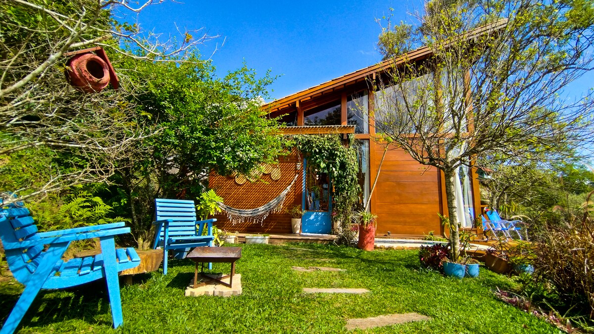 Casa de madeira com telhado inclinado e grandes janelas, cercada por gramado verde e &aacute;rvores. H&aacute; duas cadeiras azuis e uma mesa de centro no jardim, com uma rede branca pendurada na varanda. O c&eacute;u &eacute; azul claro