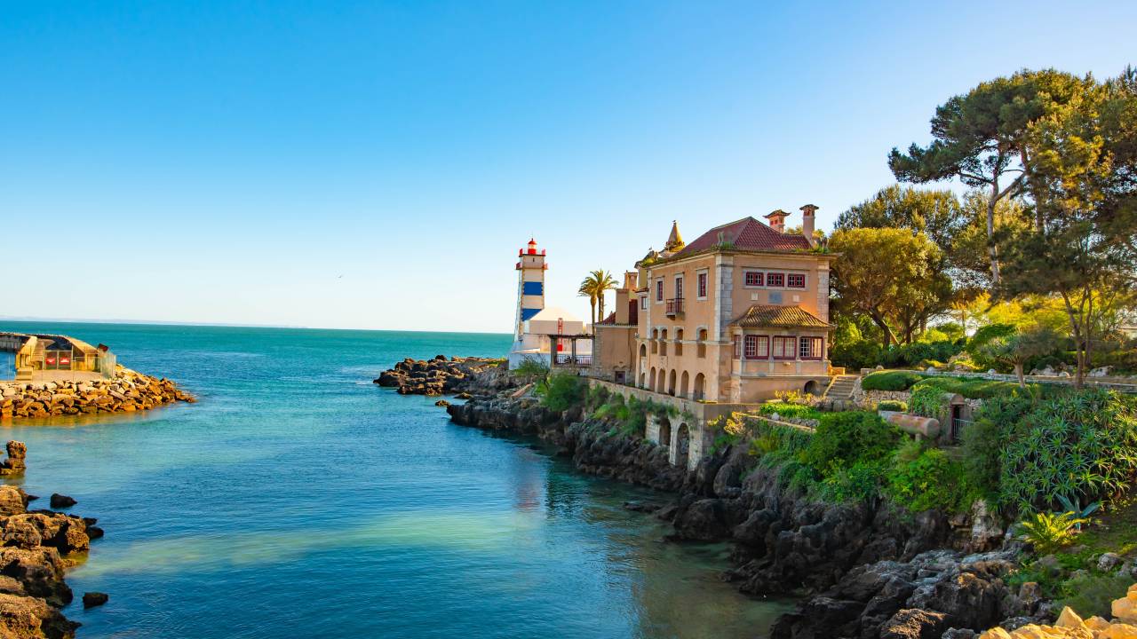 Um braço do mar de águas calmas com um casarão e um farol lá na frente