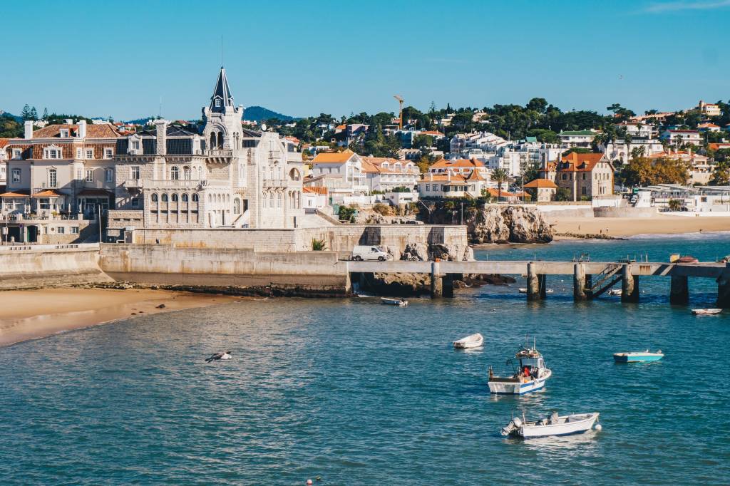 A foto mostra num primeiro plano o mar azul com barquinhos de pesca e, atrás, casas antigas do centrinho de Cascais