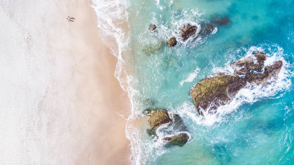 Foto aérea que mostra a areia banhada pelas águas verdes do mar, com algumas pedras