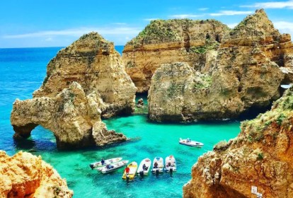 Fotografia colorida mostra barcos a flutuar num mar de águas azuis cercado por falésias e pedras que emergem da água