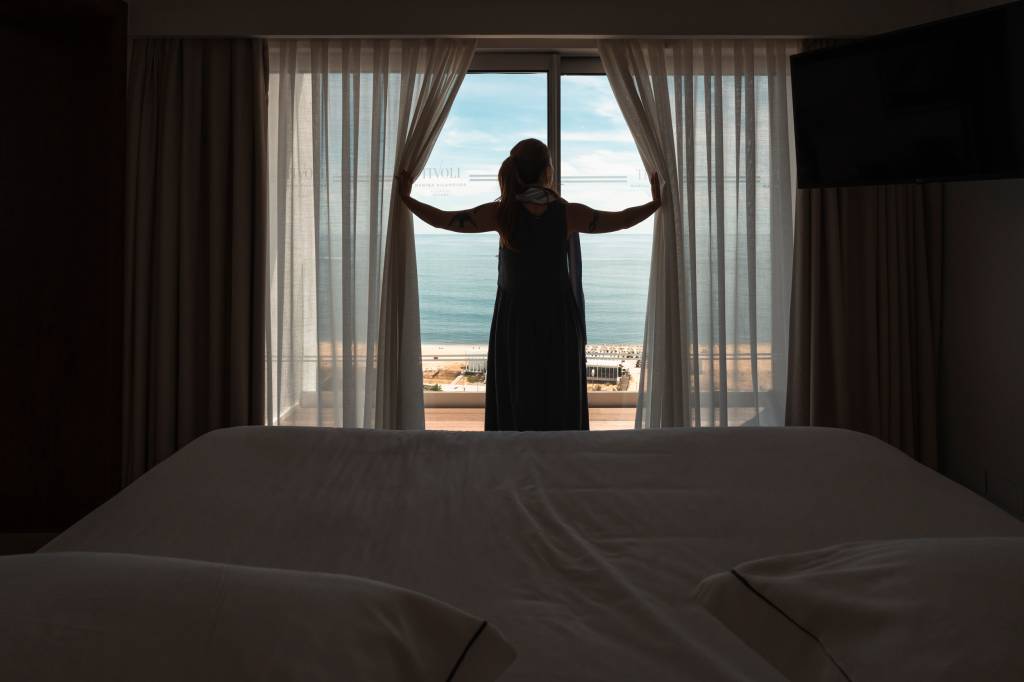 Cama do quarto do hotel, com uma mulher abrindo as cortinas, através das quais se vê o mar