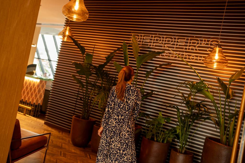 Mulher vestida de azul e branco entra no restaurante através de uma parede de madeira onde se lê Pepper's Steakhouse