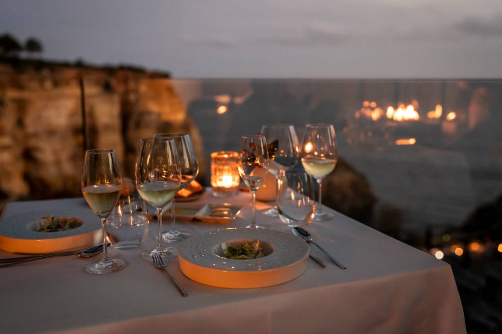 Mesa à luz de velas, com taças de vinho e pratos; ao fundo, falésia e mar