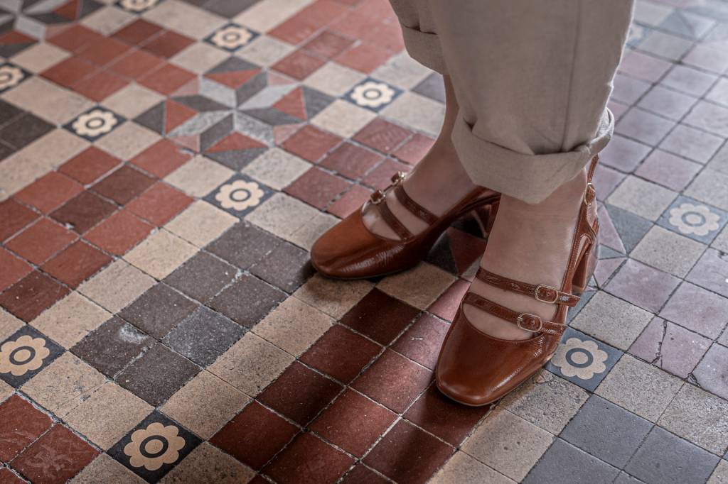 2024_Nuvem-167 Detalhe do piso em ladrilho do hotel, em tons castanhos, com os pés de uma mulher com sapatos marrom de fivelas