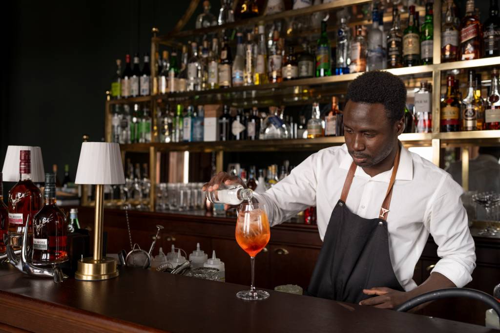 2024_Nuvem-170 À frente de uma parede repleta de garrafas de bebidas, um barman finaliza um drink de cor laranja no copo